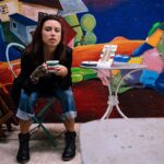 Image of a artistic young woman, enjoying a coffee in front of a colorful mural with a Sammi Italian Sandwich