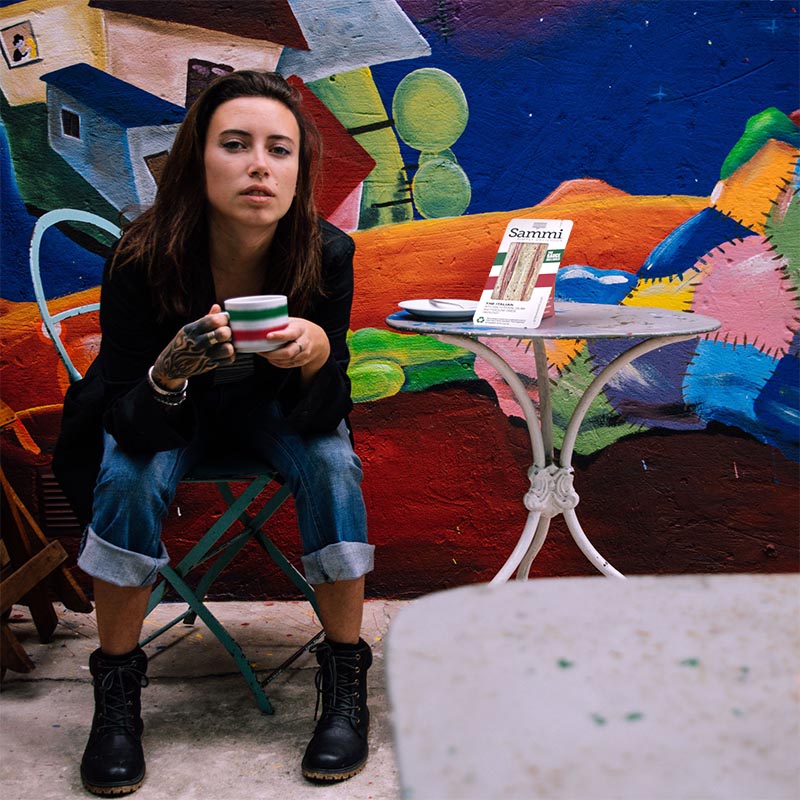 Image of a artistic young woman, enjoying a coffee in front of a colorful mural with a Sammi Italian Sandwich